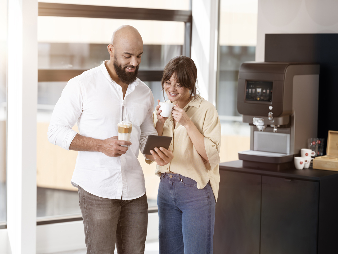 Двоє людей стоять біля кавомашини; один тримає планшет, інший — склянку з холодною кавою.